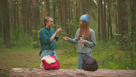 two young women enjoy a peaceful outdoor break in a lush forest, one pours a warm drink from thermos as her friend happily accepts and drinks, both sharing a moment of friendship