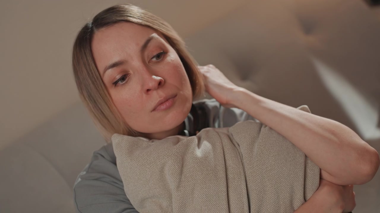 Mulher caucasiana abraçando um travesseiro no sofá, luz suave e quente do abajur, olhar cabisbaixo, segurando a almofada com ansiedade, solidão noturna, buscando conforto após um desgosto amoroso, close-up cinematográfico, vulnerabilidade emocional