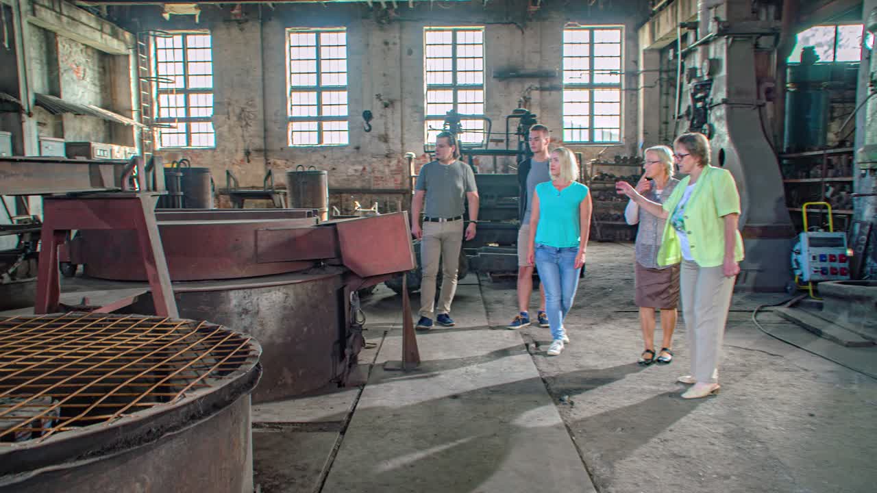 Family of four takes tour in old steel mill with industrial machines. The guide explains the gigantic machines. Slow Motion Tracking shot