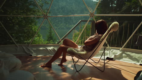 pensive woman enjoys incredible view of lake and mountains sitting in armchair with fur on glass balcony letting in suns rays. tropical holiday in modern tourist building