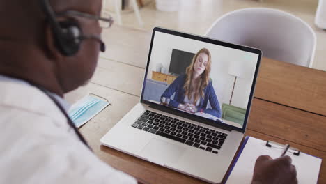 médico afroamericano que usa auriculares de teléfono tomando notas mientras hace una videollamada en una computadora portátil