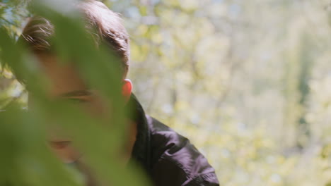 tourist kneels in forest under soft sunlight, looks thoughtfully in two directions while opening backpack to check supplies, surrounded by trees and green foliage, considering best path to follow