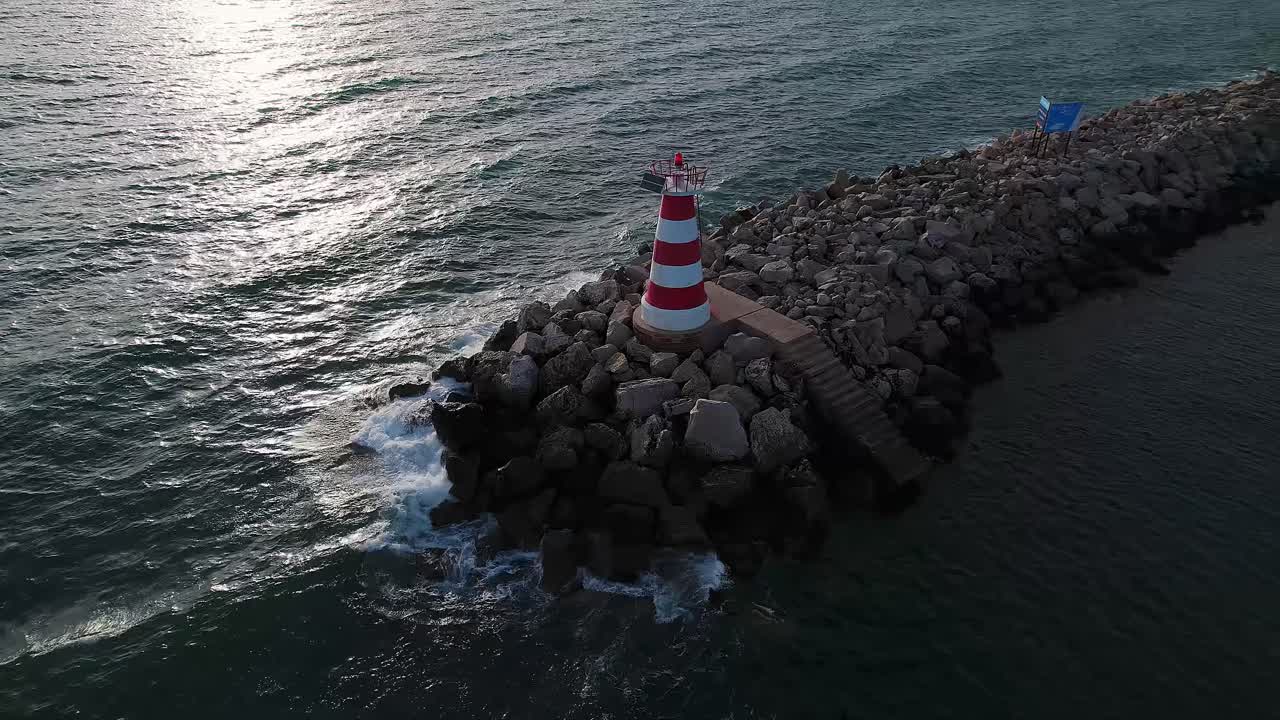 Drone orbit around the small conical Vilamoura lighthouse set on a stone jetty surrounded by Atlantic waves, marking the end of the marina entrance breakwater near Vilamoura Beach