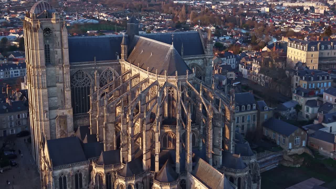 Imagem de drone circulando a nave da Catedral de Le Mans ao pôr do sol, luz dourada quente iluminando a pedra enquanto a cidade cerca o monumento e um pássaro cruza o enquadramento. França.
