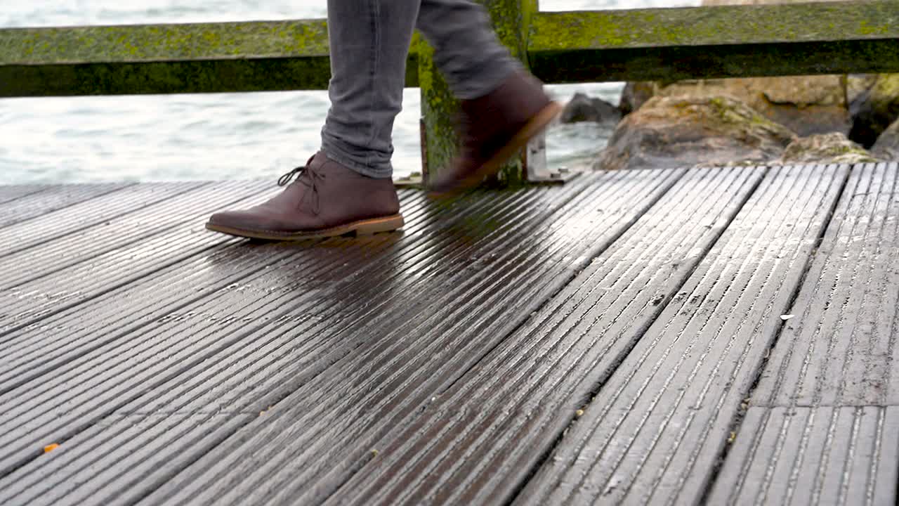 Close-up view of man's legs in jeans and brown leather boots walking on a wooden pier
