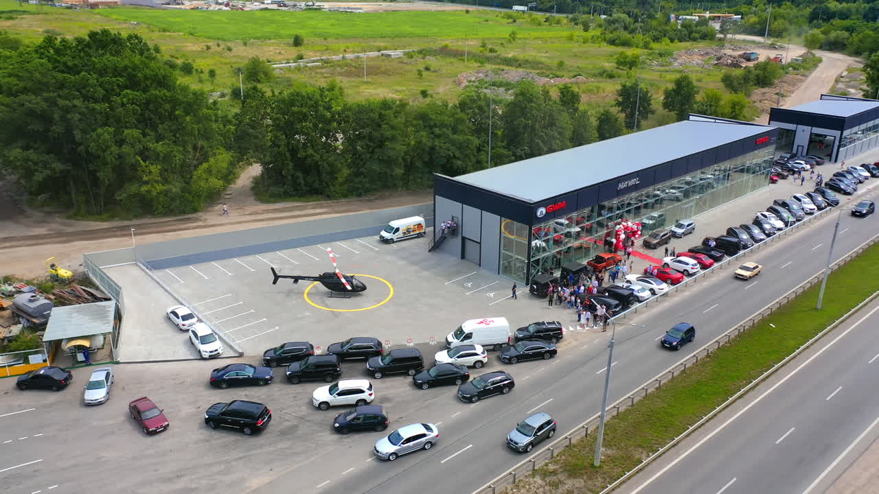Helicopter landed near the automobile centre and its bladed rotors still rotate. Car dealer customers come out to see the machine. Top view.