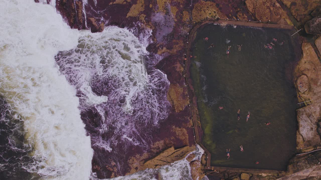 punto de vista de pájaro de nadadores en una piscina y olas frenando sobre rocas