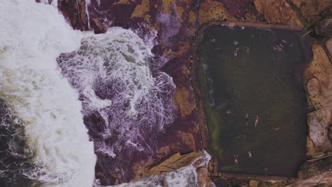 Birds-Eye-POV-of-swimmers-in-a-pool-and-waves-brakeing-over-rocks