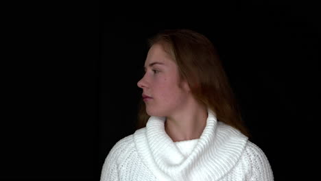 a close-up of a woman wearing a white knitted jumper, turning her head to the side, on a seamless black studio background