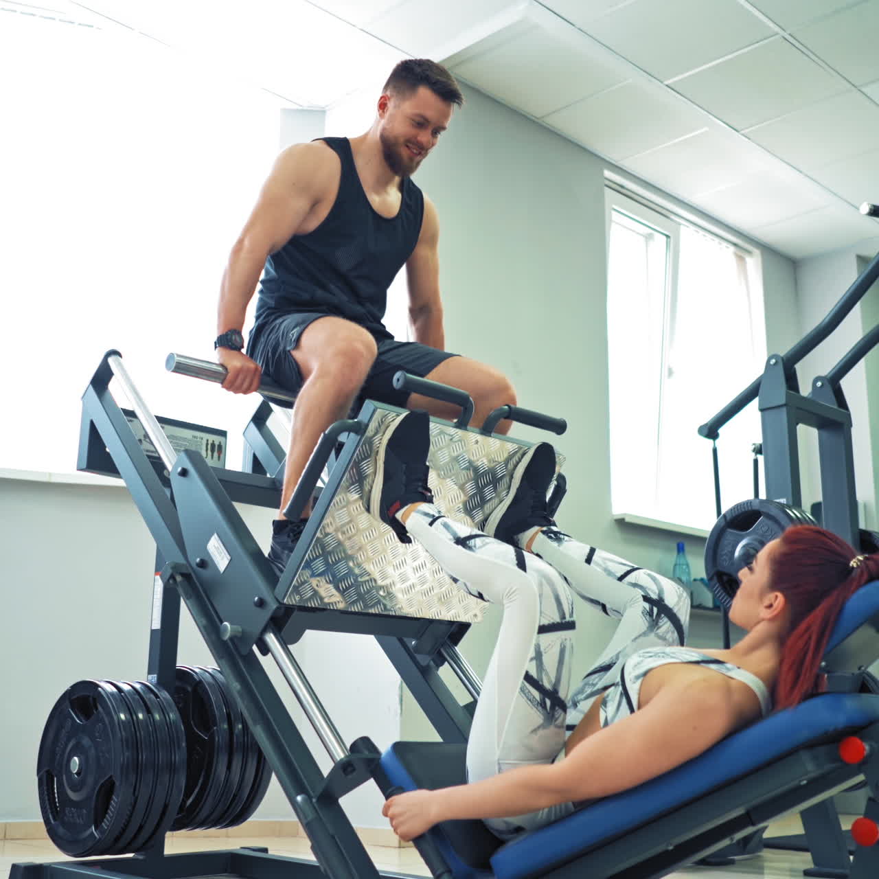 Sporty girl doing exercise for legs on a modern simulator in the gym. Woman is lifting man by her strong legs on a fitness machine in the light sports center.