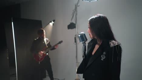 focused female vocalist in black performing with a vintage microphone, guitarist in background, against a white studio backdrop