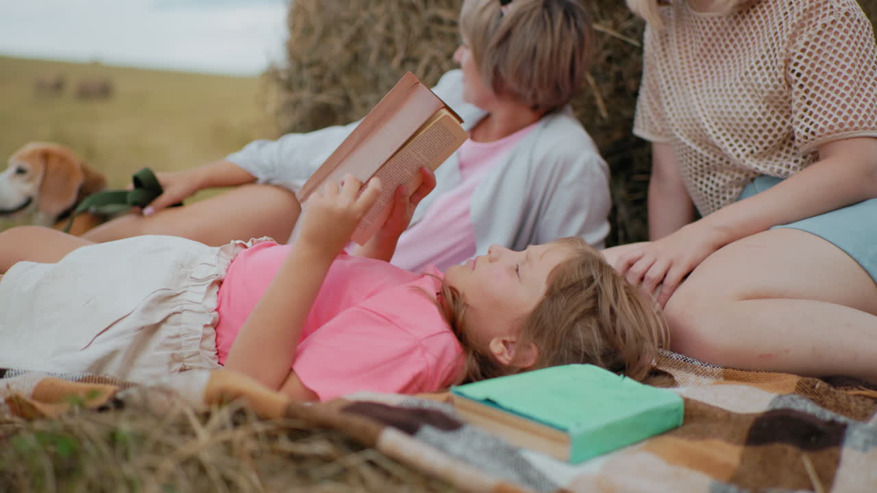 Close-up of little girl in pink shirt reading her book while lying on picnic blanket, green book near head, family relaxing with dog on leash and enjoying peaceful time outdoors in the countryside