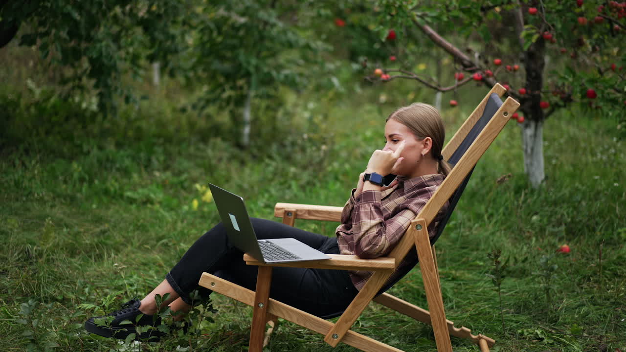 Busy young lady speaks on the phone looking at laptop in front of her. Blonde woman freelancer working remotely in her garden.