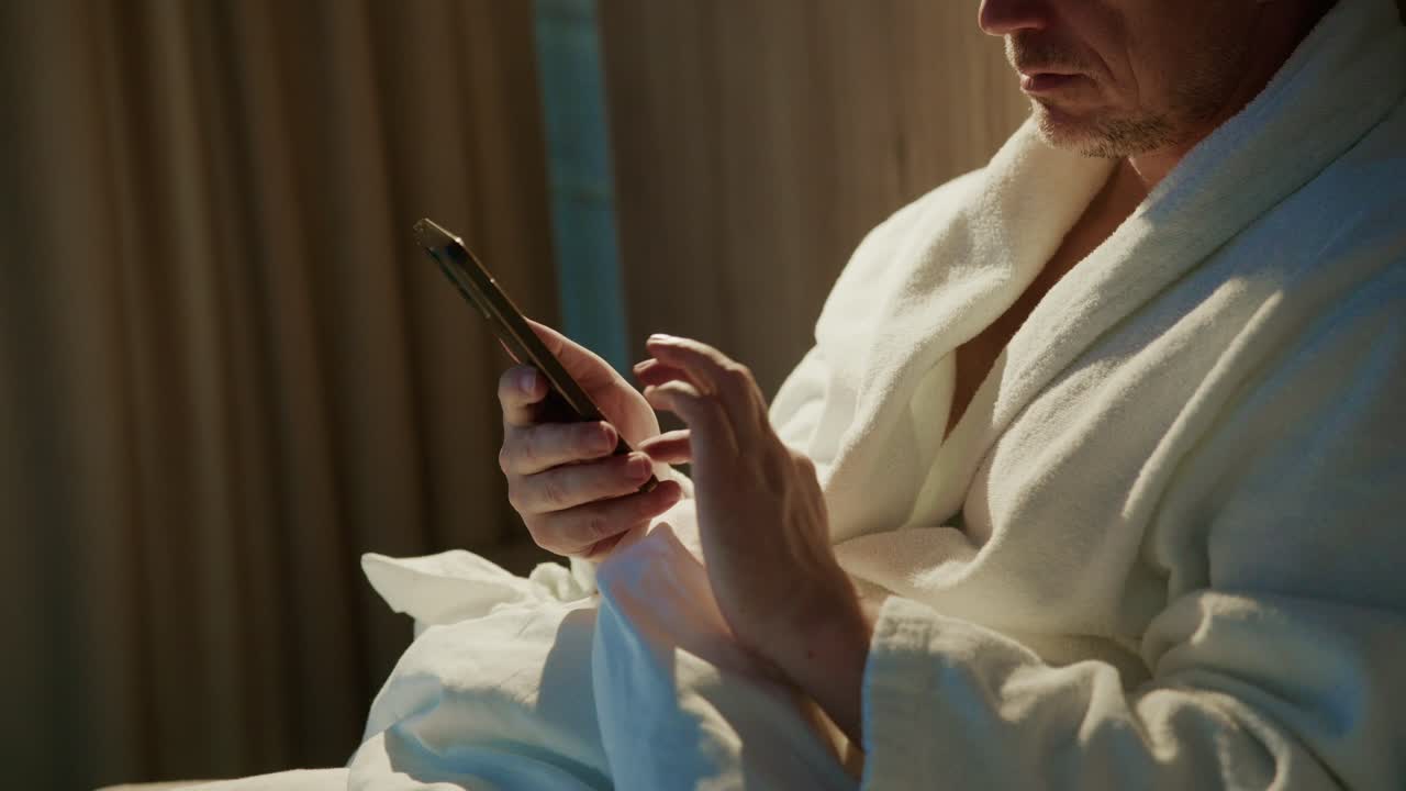 Muž v měkkém županu sedí v tlumené místnosti a soustředí se na obrazovku svého chytrého telefonu. Klidná atmosféra naznačuje, že si užívá osobní chvíle a připravuje se na večerní odpočinek.