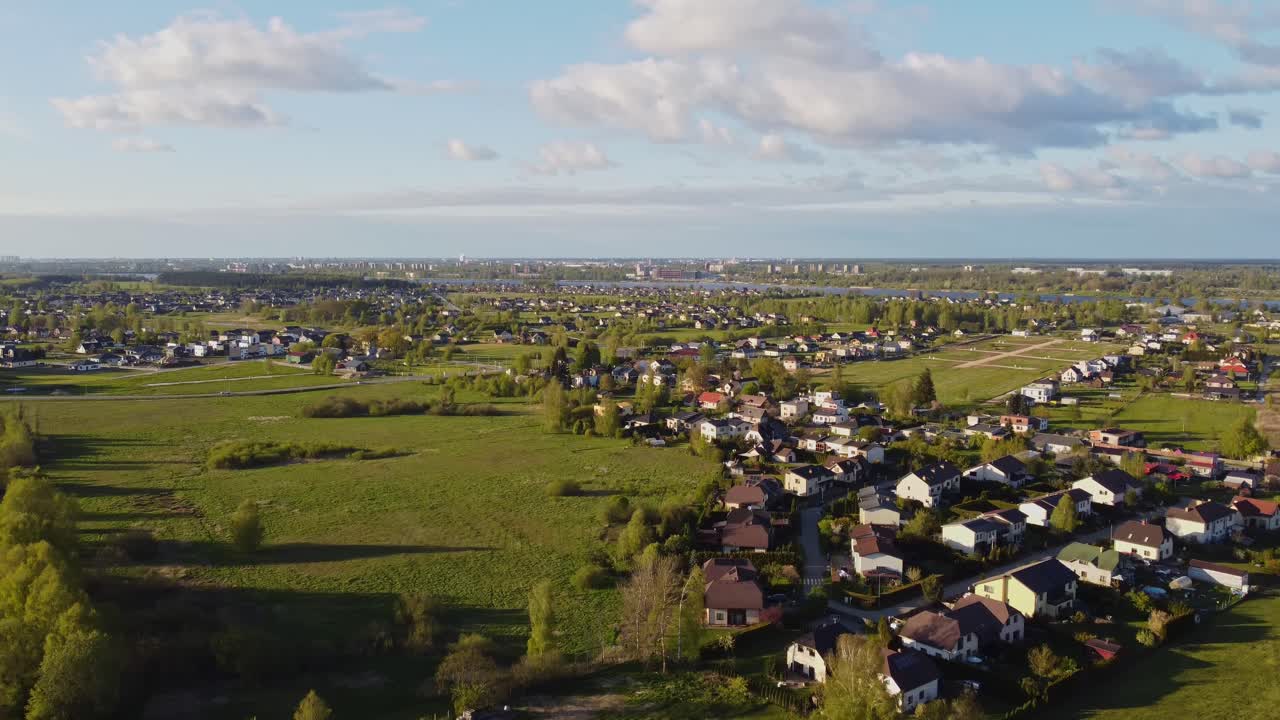 Drone slowly pulls away above Katlakalns residential area showing houses, spring green fields, tree lines, and Riga skyline on the horizon under scattered clouds and afternoon light