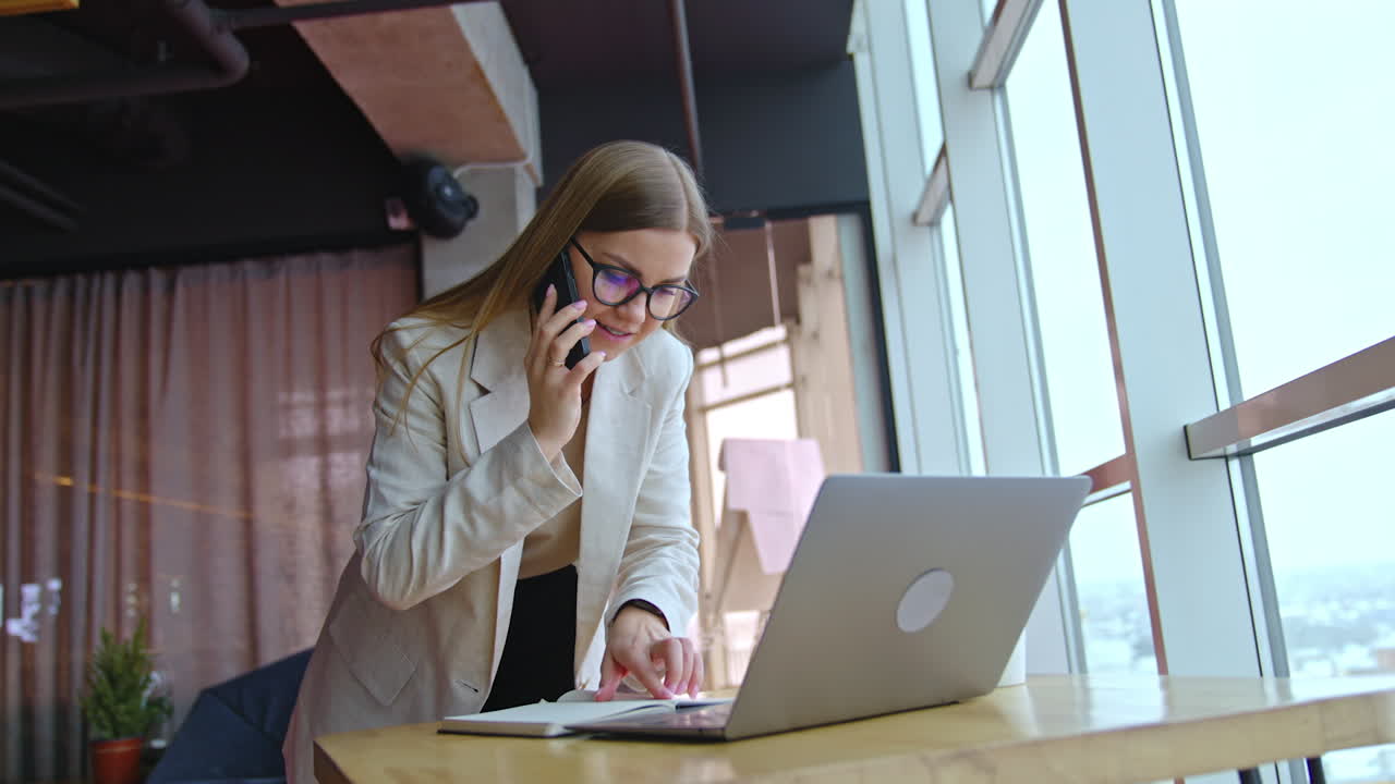 Beautiful working lady standing at the desk with laptop. Busy woman speaking on the phone and checking her notes. Office background.