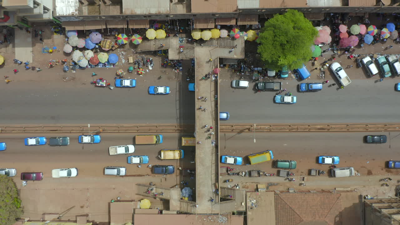A striking, high-angle drone shot directly over a busy street in Guiné-Bissau, West Africa. Features an overpass, traffic congestion, and colorful market stalls