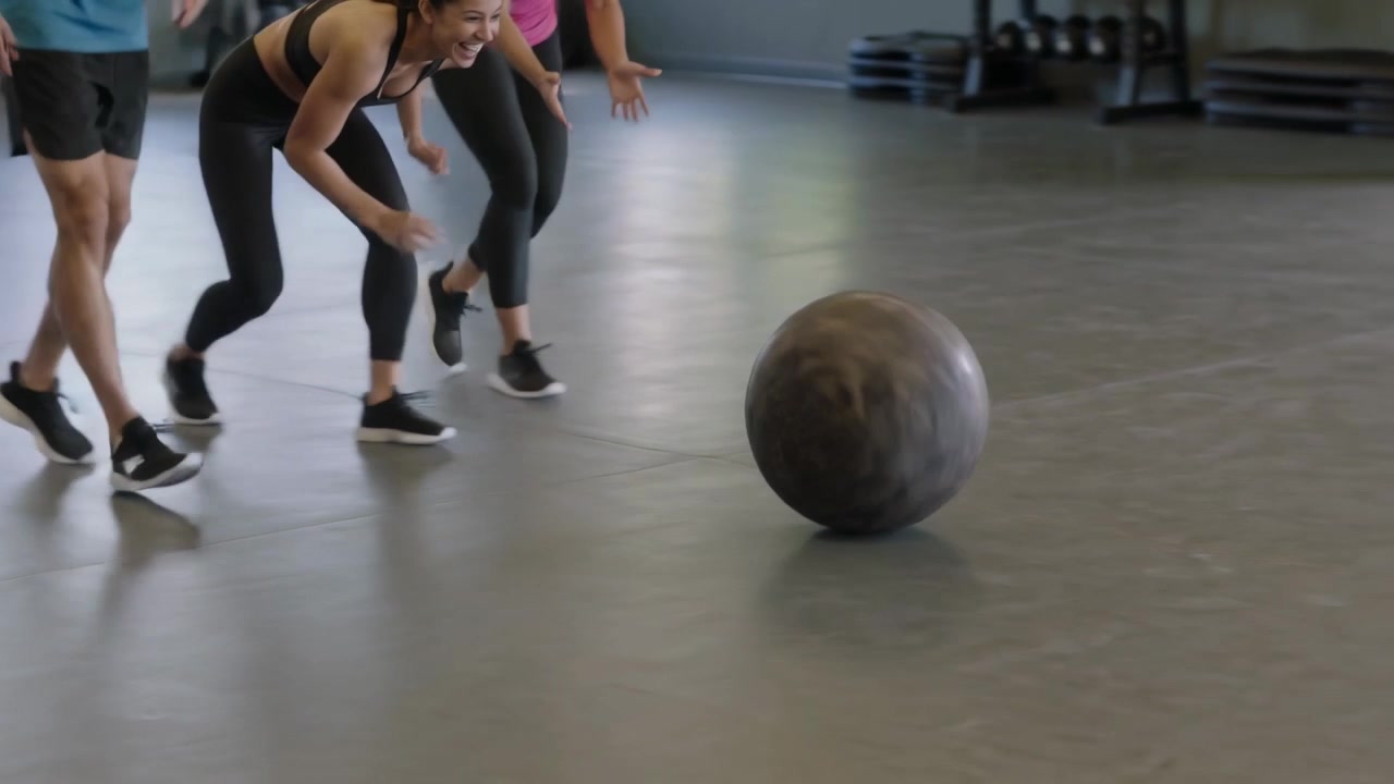 Reacting to medicine ball rolling, trio in sportswear stepping across gym floor to retrieve ball