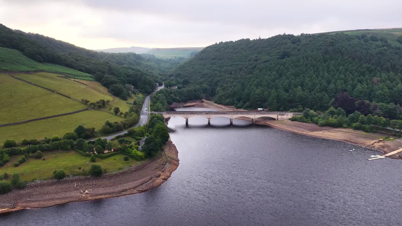 Drone footage glides above a multi-arched bridge spanning a wide reservoir, surrounded by lush countryside and dense forest under overcast daylight