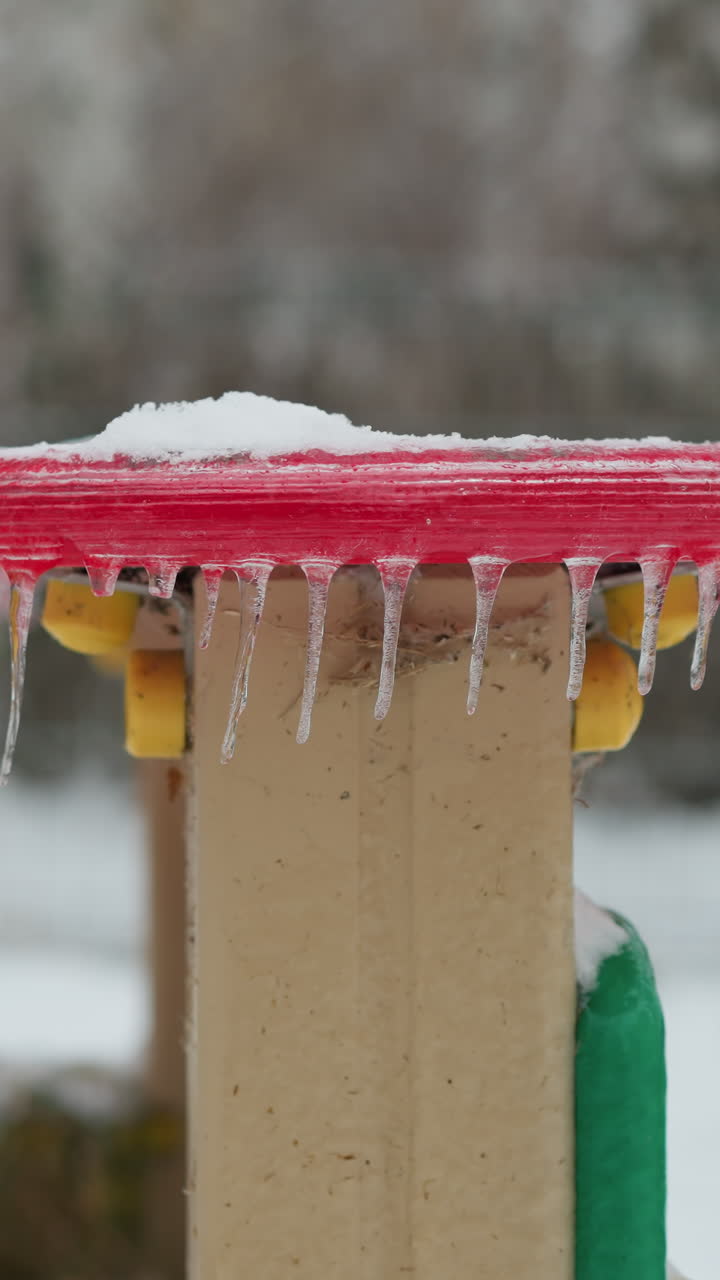 놀이터 구조에 매달린 얼음 조각과 함께 은 원형 얼음 표면의 클로즈업, 얼음 세부 사항의 대조와 흐릿한 눈 바탕을 포착, 멀리 자동차의 흐릿한 시야