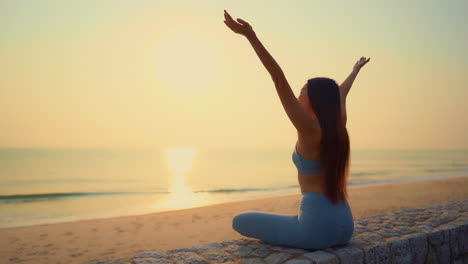 Vista-Trasera-De-Una-Joven-Asiática-Haciendo-Meditación-Junto-Al-Mar,-Playa,-Océano-Al-Amanecer,-Brazos-Abiertos-Saludando-Al-Sol