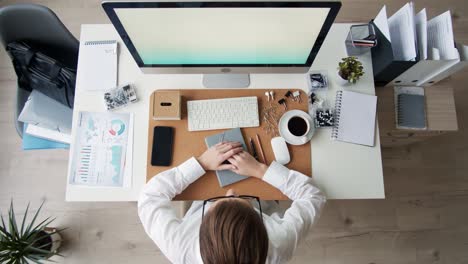 flat lay of young businesswoman wearing white shirt and glasses sitting at desktop at pc screen, putting on wireless headphones and answering video call from business partner