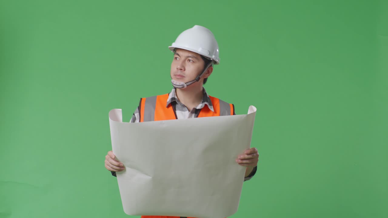 ingeniero masculino asiático con casco de seguridad mirando el plano en sus manos y mirando a su alrededor mientras camina en el estudio de fondo de la pantalla verde