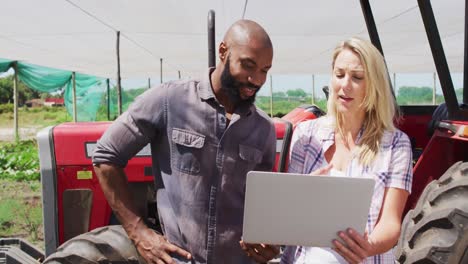 video of happy diverse woman and man using laptop and talking in front of tractor