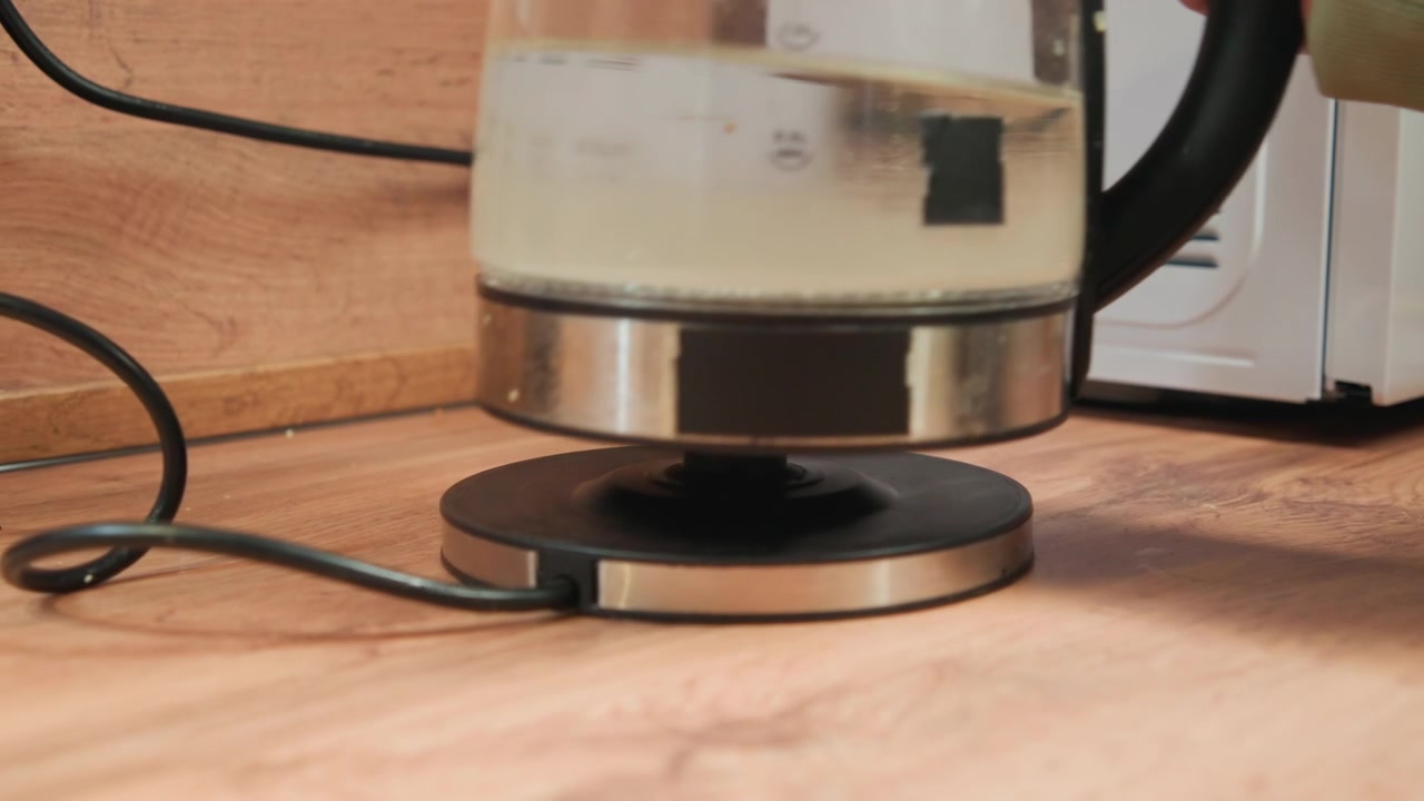 Transparent electric kettle sits on round hotplate atop wooden countertop with braided cord connected, water bubbling inside, steam rising and warm light reflecting across glass and metal surfaces