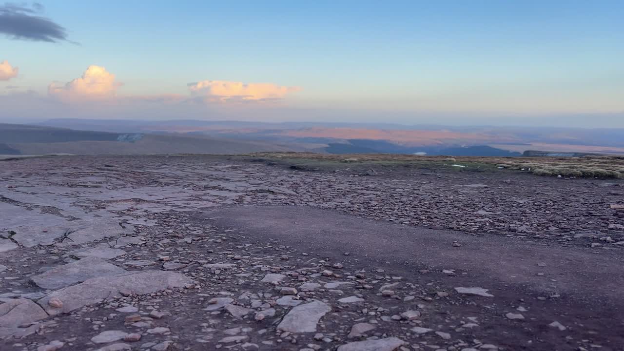 Altopiano sommitale di pietra fessurata sul Cribyn con ampie vedute sulle Brecon Beacons sotto una morbida luce serale e nuvole sparse che creano un paesaggio montano calmo ed esteso.
