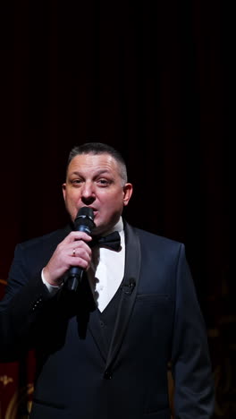 speaker on stage. male host in elegant costume speaking into a microphone looking on camera on the background of red curtains in the theater. slow motion. vertical video