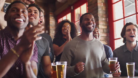 Grupo-De-Amigos-Y-Amigas-Celebrando-Mientras-Ven-El-Juego-En-La-Pantalla-En-El-Bar-Deportivo