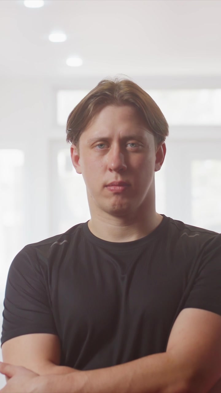 Young man stands in bright indoor space with large windows wearing. His arms are initially clasped at waist level before crossing. Scene is well lit by natural daylight streaming