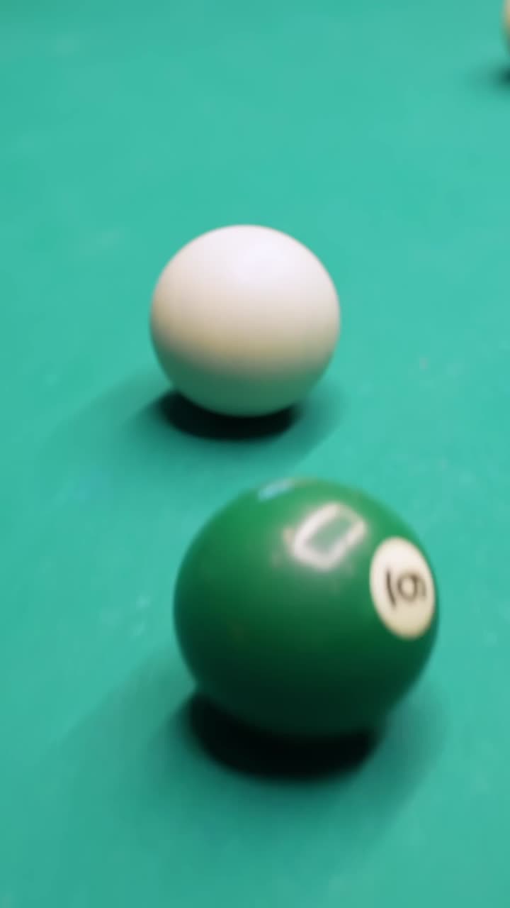Vertical close-up of a billiard cue striking the cue ball to hit a green ball on green felt, shallow focus for sports bar, competition, and game night b-roll