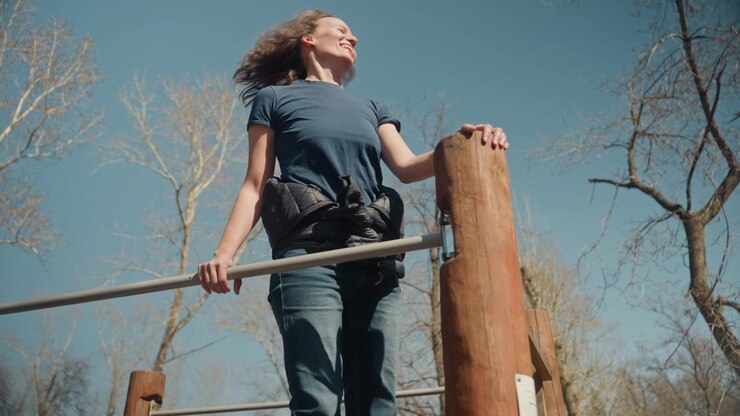 Close up of woman performing outdoor exercise on wooden equipment holding onto metal bars and wood posts. Woman wears casual clothing with item around her waist as woman moves