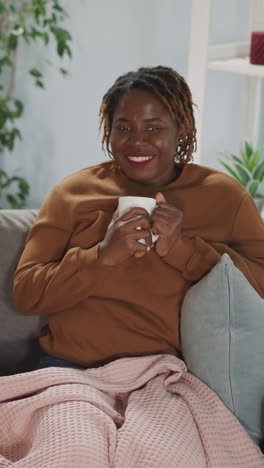 multinational friends enjoy watching interesting tv program with cup of hot drink. indian and african american women rest on couch smiling and talking