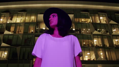 woman in hat under purple light looking from side to side with futuristic building in background - low-angle shot