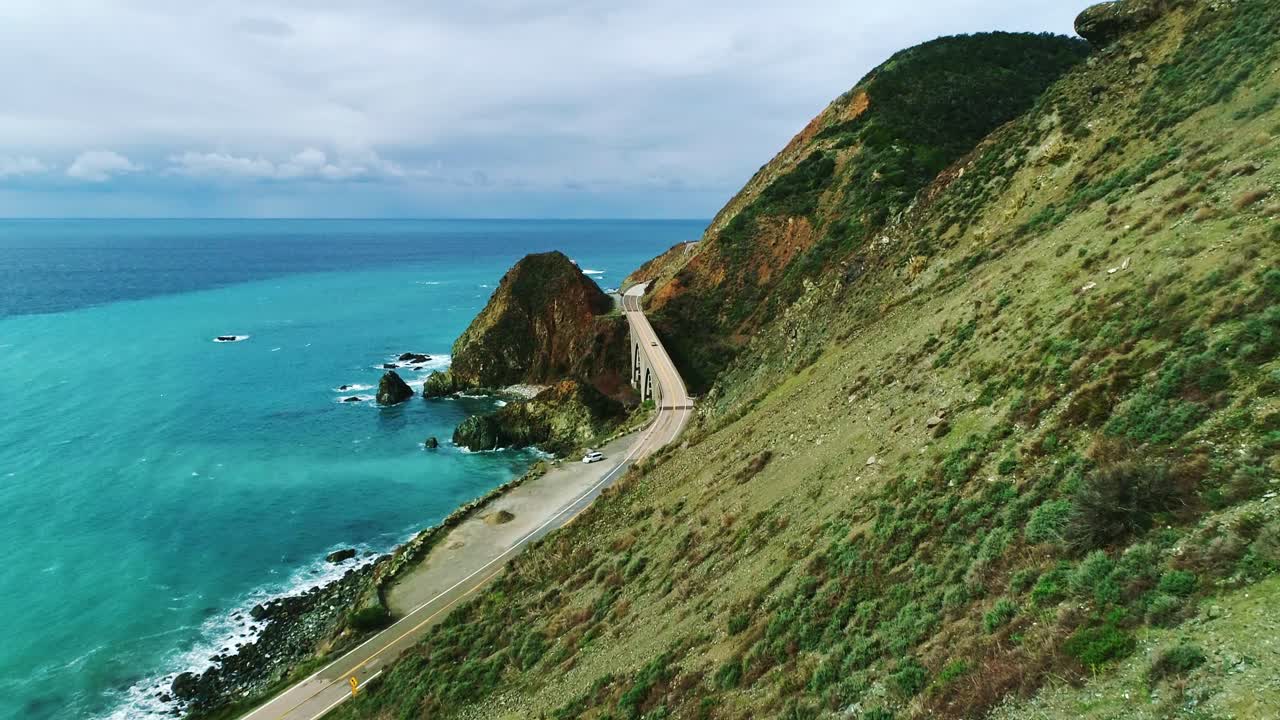 Coastal road winds along steep cliffs above vibrant turquoise Pacific Ocean surf