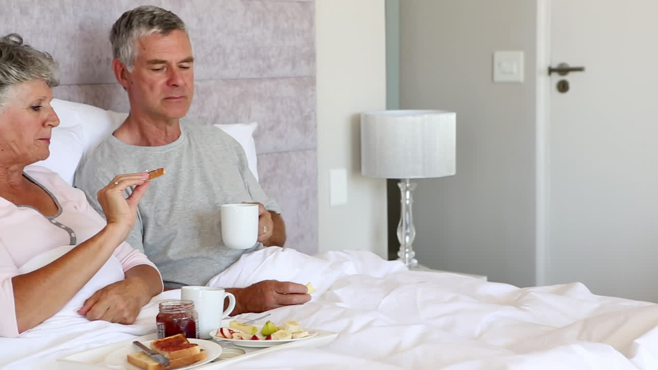 성숙 한 커플 이 침대 에서 아침 식사 를 하고 있다