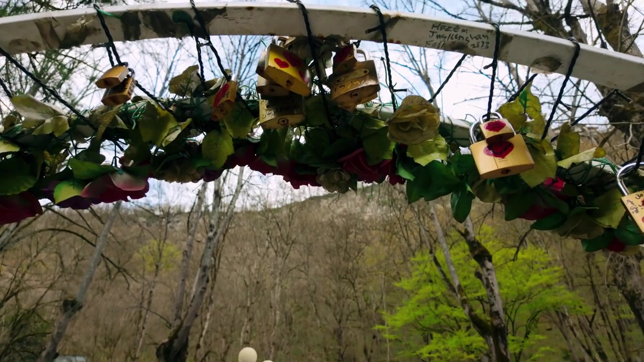 Arco decorativo adornado con muchos 'candados del amor' coloridos y hojas verdes, ubicado en un parque con árboles desnudos y follaje fresco de primavera, simbolizando afecto y compromiso en un entorno natural