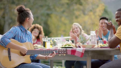 Mujer-Birracial-Tocando-La-Guitarra-Con-Un-Grupo-Diverso-De-Amigos-En-La-Mesa-En-El-Jardín,-Cámara-Lenta