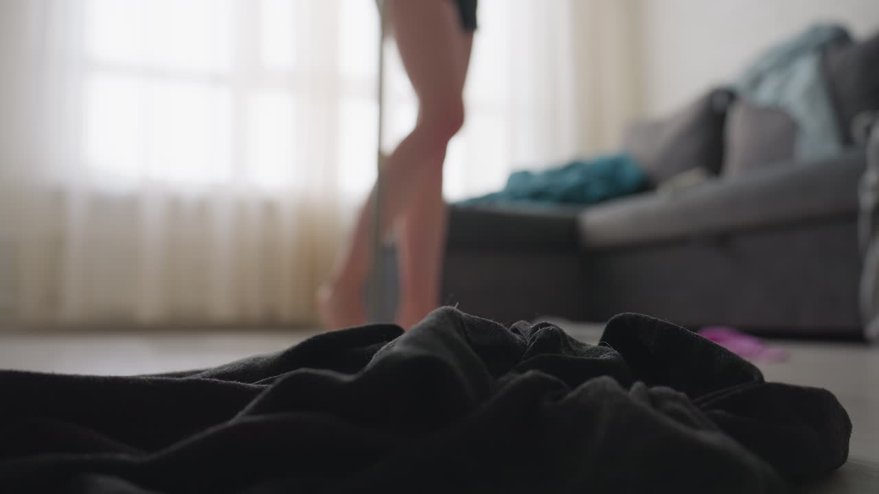 Close up of black cloth on floor with blurred view of woman in shorts holding mop walking past messy living room, sofa covered with scattered clothes and blanket in background