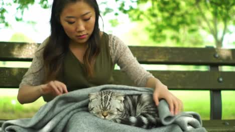 a woman comfortably cuddles her cat in a cozy blanket while relaxing on a park bench surrounded by lush greenery under a clear blue sky