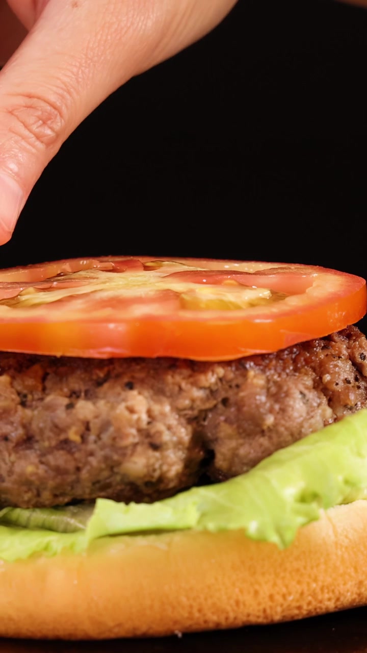Una mano aggiunge delicatamente una fetta di pomodoro su un hamburger di manzo con lattuga e panino, ripreso in primo piano con illuminazione da studio brillante e uno sfondo nero.