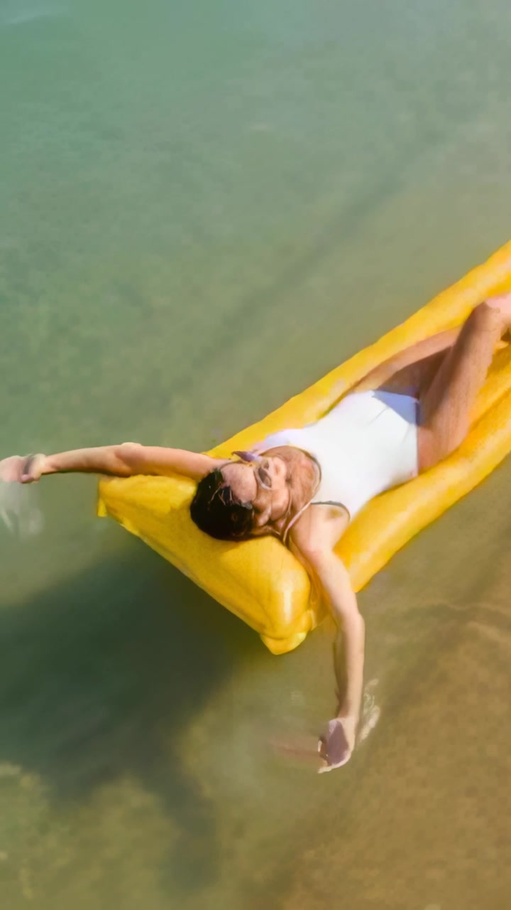Vertical video: Extending woman floating on yellow float at beach with white swimsuit and shades