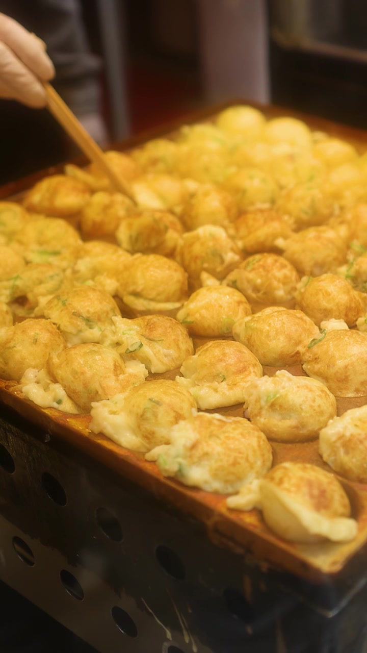 Primo piano di uno chef che usa spiedini per girare palline di takoyaki dorate su una griglia.