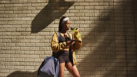 african american woman exercising outdoors drinking water and using smartphone in the city