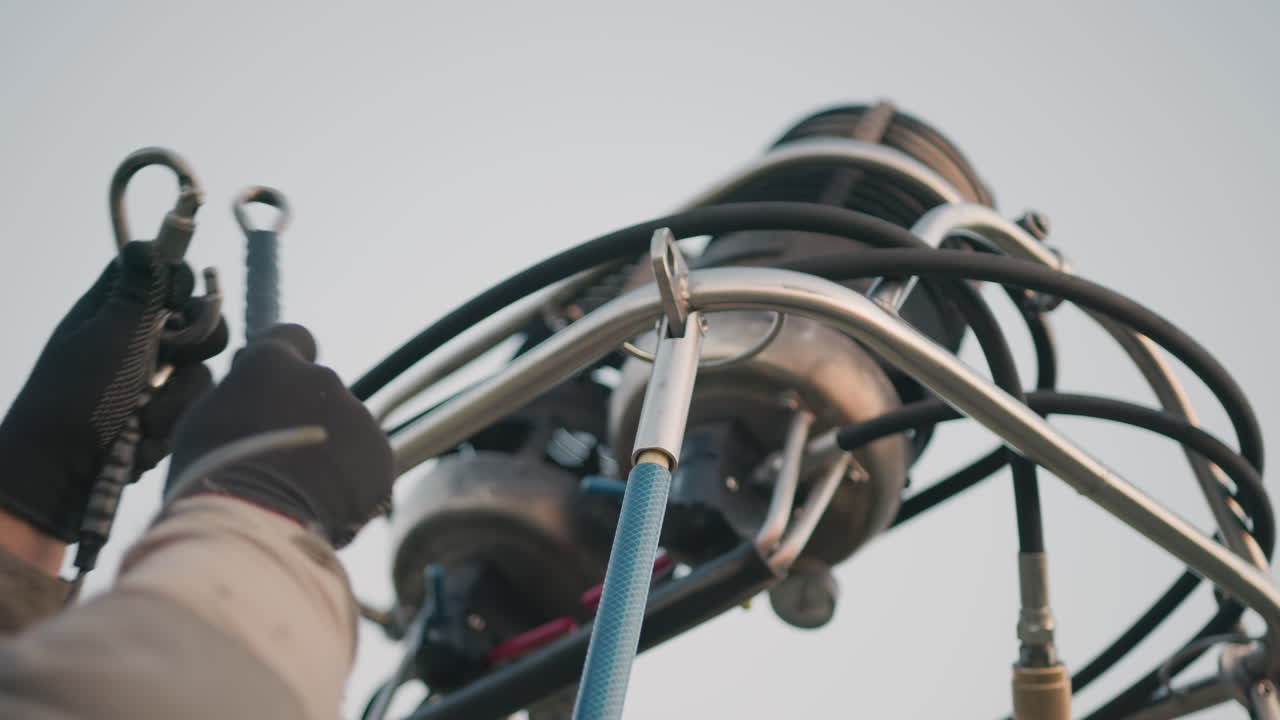 close up of gloved hands adjusting metal burner system of hot air balloon showing detailed mechanical parts including valves, tubes, and braided lines with clear sky background