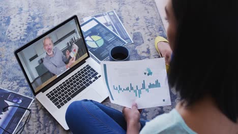 Mixed-race-businesswoman-sitting-on-floor-using-laptop-having-video-call-with-male-colleague