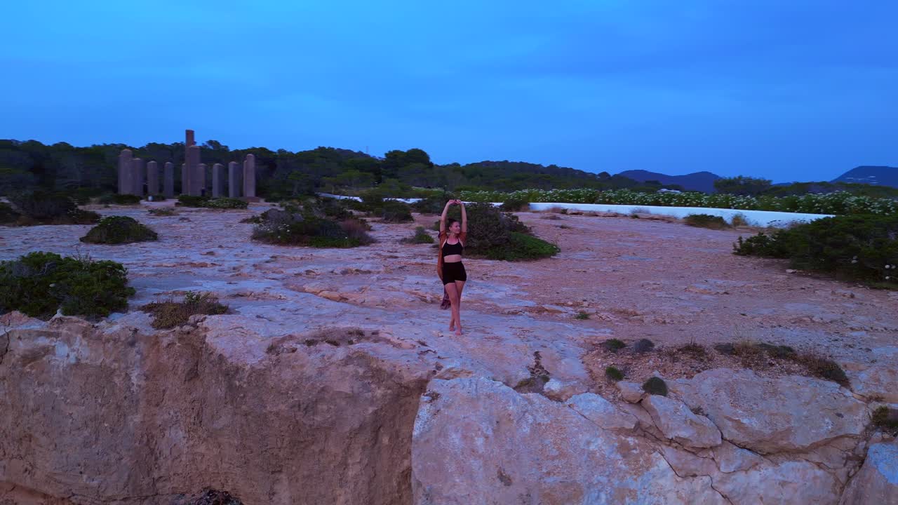 Woman performing a yoga tree pose on a rocky cliff during a serene sunset in Cala Llentia Ibiza. Fabulous aerial view flight circle drone footage